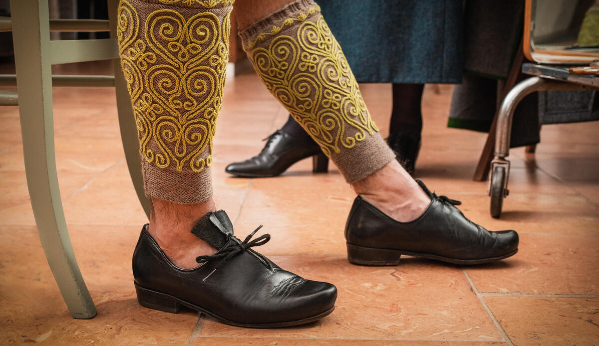 The photo shows two men's shins adorned with grey woollen socks (so-called "Loferln"). The socks are embroidered with yellow thread in scrollwork. Black Haferl shoes are worn with them.