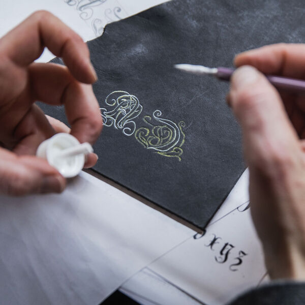 Close-up of needlework: The right hand holds a brush, the left hand holds a white paint container. On the table is a piece of brown leather on which two ornamental initials are being pre-drawn to be subsequently embroidered with the quill.