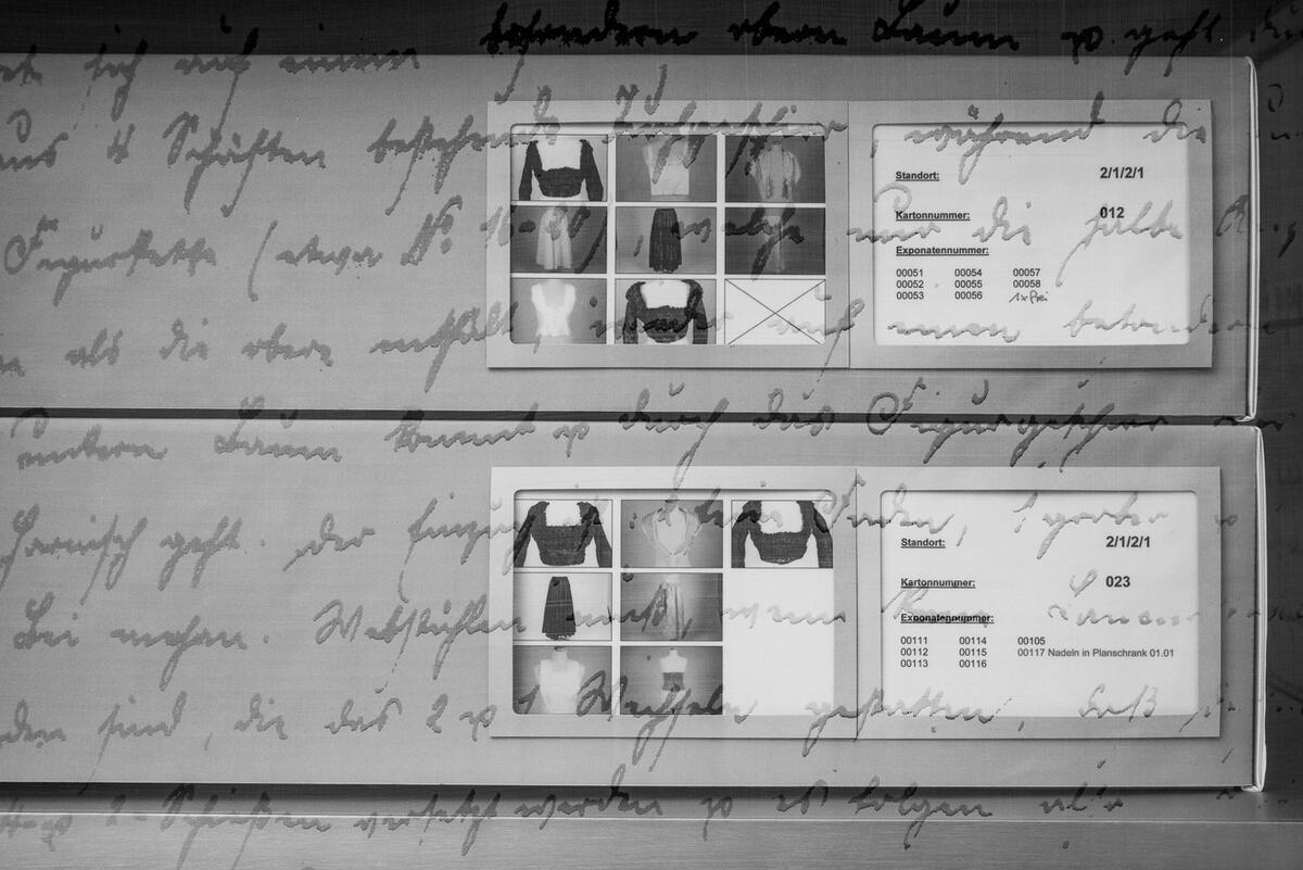 Detailansicht von gestapelten Archivschachteln aus grauem Karton. Miniaturfotos und Beschriftungen darauf verweisen auf den Inhalt.