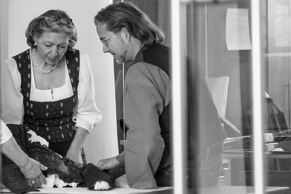 Two employees of the Centre for Traditional Costume examine a historical waistcoat that is spread out on a table in front of them. The employee (left in the picture) is wearing a patterned traditional dress and a white dirndl blouse underneath; opposite her, in profile view, you can see the upper body of her colleague, who is dressed in a shirt and waistcoat.