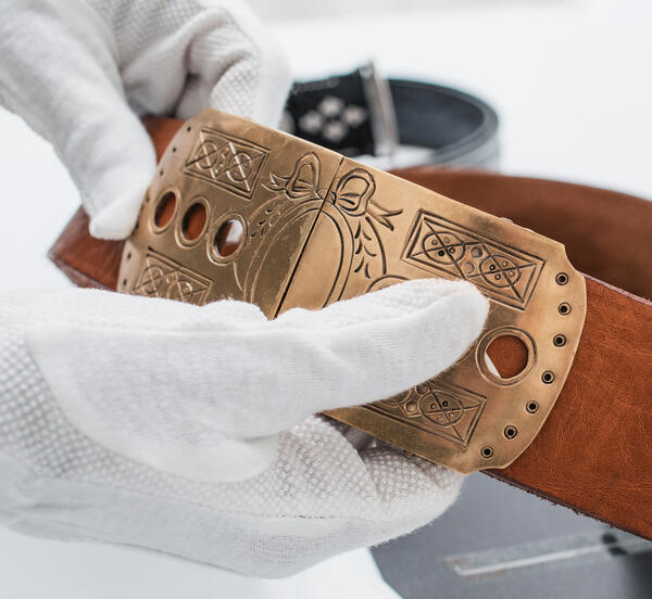 The photo shows a medium brown leather belt with an ornate bronze buckle presented by two hands in white gloves.