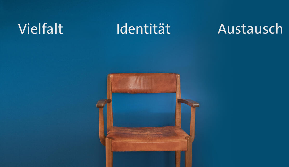 A simple antique wooden chair with a brown leather seat and armrests stands in front of a blue wall of the Centre for Traditional Costume. The wall behind it reads in white letters: "Diversity", "Identity" and "Exchange" - three words from a longer wall print.
