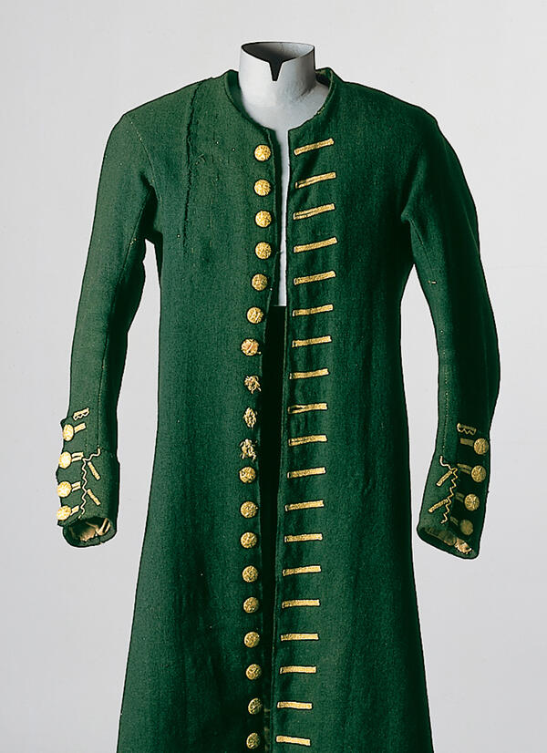 A dress-up doll presents a "Justacorps" in the colour forest green with golden buttons and decorated buttonholes. The garment for men origins from Lenggries, and is dated around 1810.