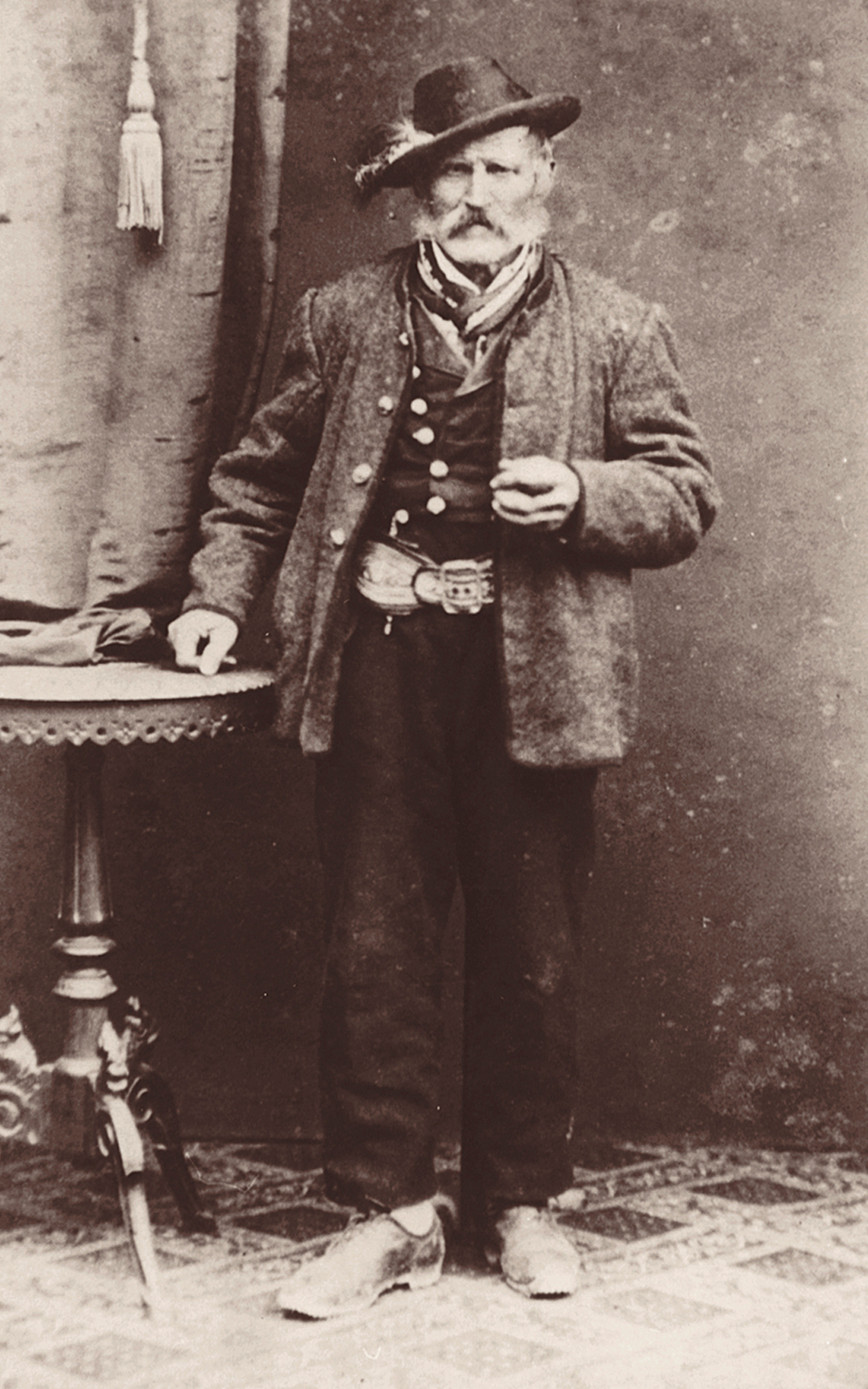 Die Schwarz-Weiß-Photographie, datiert um 1875, zeigt einen älteren Bauern in Tracht aus dem oberbayerischen Jachenau. Er posiert neben einem Beistelltisch mit drapiertem Vorhang.