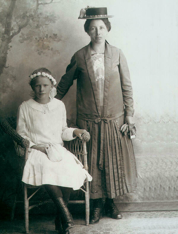 Die Schwarz-Weiß-Photographie aus Attenberg um 1925/30 zeigt ein Mädchen in ihrem Firmkleid, das auf einem Korbstuhl sitzend neben ihrer festlich gekleideten Mutter in die Kamera lächelt. Hinter den beiden ist eine Landschaftstapete zu sehen.