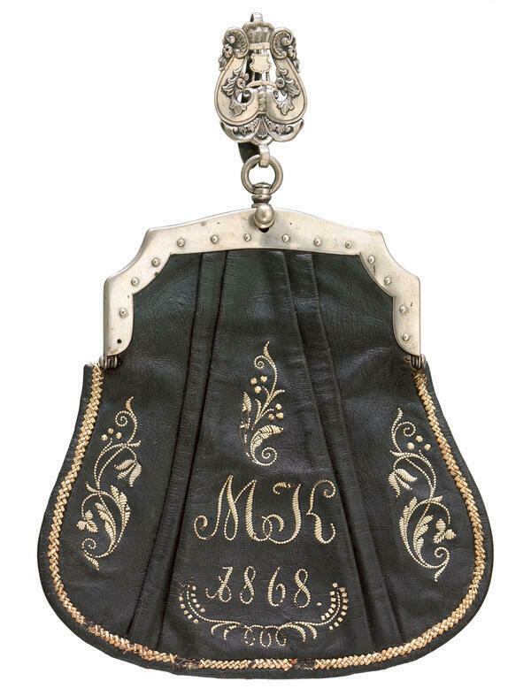 Die oberbayerische Kellnerinnen-Tasche aus dem Jahr 1868 ist aus schwarzem Leder gefertigt. Die Vorderseite zeigt ein Monogramm mit den Initialen MK, das ebenso wie die Jahreszahl und Schmuckmuster mit dem Federkiel aufgestickt wurden. Schließe und Aufhängung der Tasche sind aus versilbertem Messing.