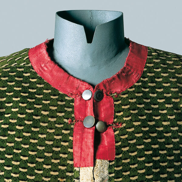 Die Kleiderpuppe trägt ein grün gemustertes Leinen-Miederleibchen aus dem Isarwinkel um 1800. Kragen und Ausschnitt sind in rotem Seidensamt gefertigt, verschlossen von zwei Reihen Metallknöpfen.