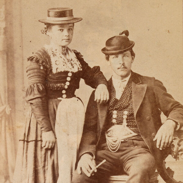 Die Sepia-Photopgrahie zeigt ein junges Bauernpaar aus dem Rupertiwinkel um 1875 in festlicher Tracht. Der Mann sitzt rechts neben der Frau, deren Arm auf seiner Schulter ruht.