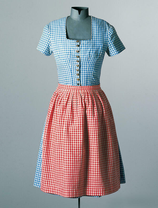 The dress-doll wears a simple cotton dirndl dress from Groweil around 1955. The short-sleeved dress is blue and white, the apron is red and white plaid. Ten metal buttons close the bodice.