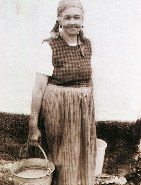 The black-and-white photograph shows a smiling older peasant woman from Rehling around 1950/60. She is wearing a patterned work dress, a short-sleeved white blouse underneath, a headscarf and holds a bucket in each hand.