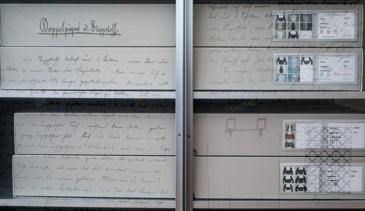 Detailansicht der vorne mit ihren Inhalten beschrifteten Aufbewahrungsschachteln aus grauer Pappe. Gelagert sind diese in den Depot-Regalen des Zentrums für Trachtengewand des Bezirks Oberbayern.