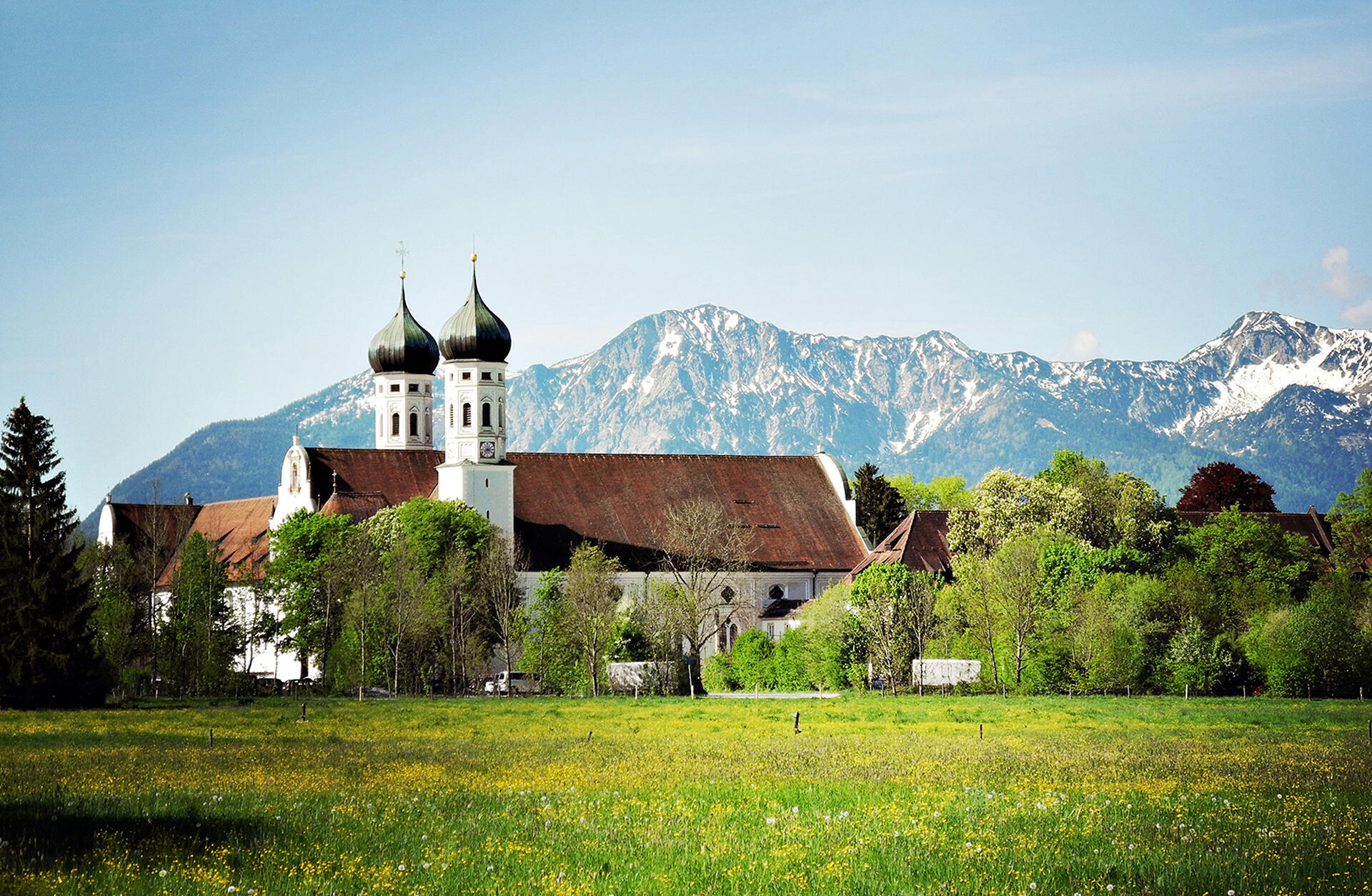 Blick auf die Anlage des Klosters Benediktbeuern mit seinen beiden Zwiebeltürmen vor einer sommerlichen Alpenkulisse.