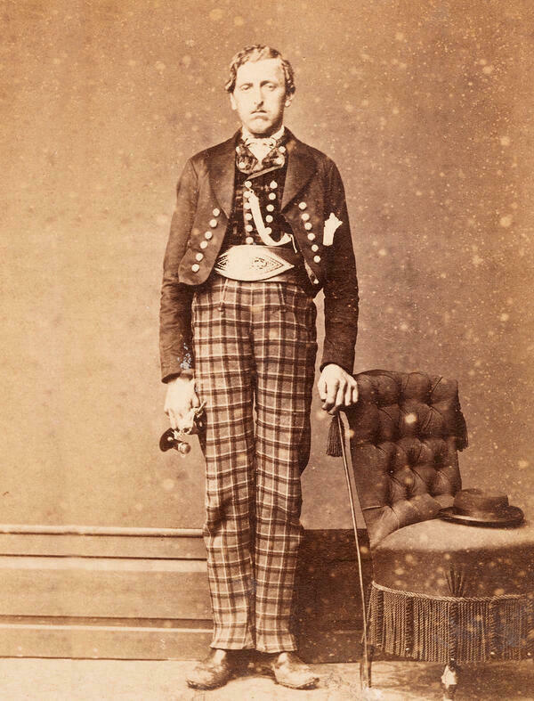 The sepia photograph shows a farmer from Upper Austria about 1870, standing and looking into the camera with a serious expression. He wears a jacket and underneath a waistcoat, both decorated with silver buttons, a wide leather belt with quill stitching, as well as checked trousers and leather shoes. His right hand rests on the back of a padded chair, on the seat of which his hat is placed.